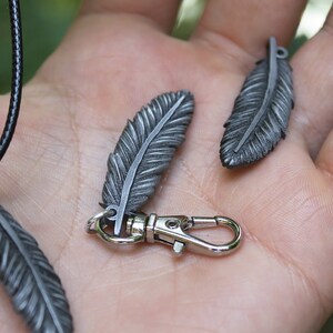 Op de afbeelding: Close-up van verschillende zilverkleurige veervormige hangers. E&eacute;n is bevestigd aan een zwarte koordketting, een andere aan een zilveren sluiting en een derde is los. De veren hebben gedetailleerde aderen.