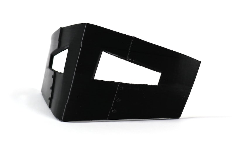May include: Black cardboard mask with two eye holes and a small opening on the top. The mask has a textured surface and is designed to cover the eyes and nose.