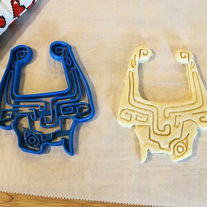 May include: Two cookie cutters in the shape of a video game character's mask. One cutter is blue plastic and the other is a light brown cookie dough.