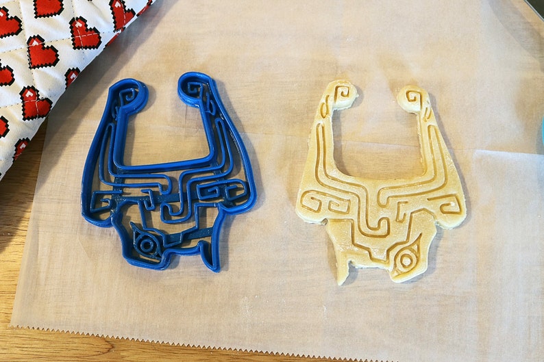 May include: Two cookie cutters in the shape of a video game character's mask. One cutter is blue plastic and the other is a light brown cookie dough.