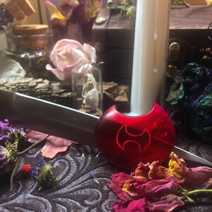 May include: A red, plastic, decorative object with a stylized symbol resembling a double-headed axe. The object is on a dark, textured surface with dried flowers and other decorative items.