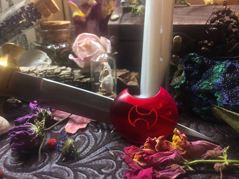 May include: A red, plastic, decorative object with a stylized symbol resembling a double-headed axe. The object is on a dark, textured surface with dried flowers and other decorative items.