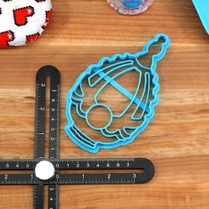 May include: A blue plastic cookie cutter shaped like a baby carriage. The cutter is on a wooden surface.