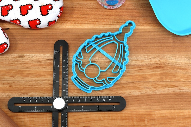 May include: A blue plastic cookie cutter shaped like a baby carriage. The cutter is on a wooden surface.