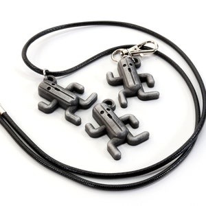 May include: Three silver-colored pendants shaped like stick figures with arms and legs, hanging from a black cord. The pendants are attached to a silver-colored clasp.