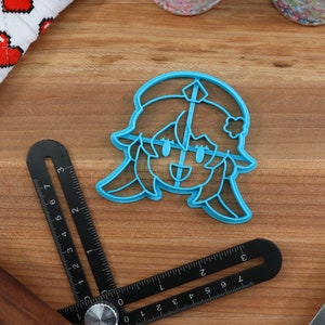 May include: A blue plastic cookie cutter shaped like a cartoon character with a flower in their hair. The character has a smiling face with closed eyes and a small mouth.