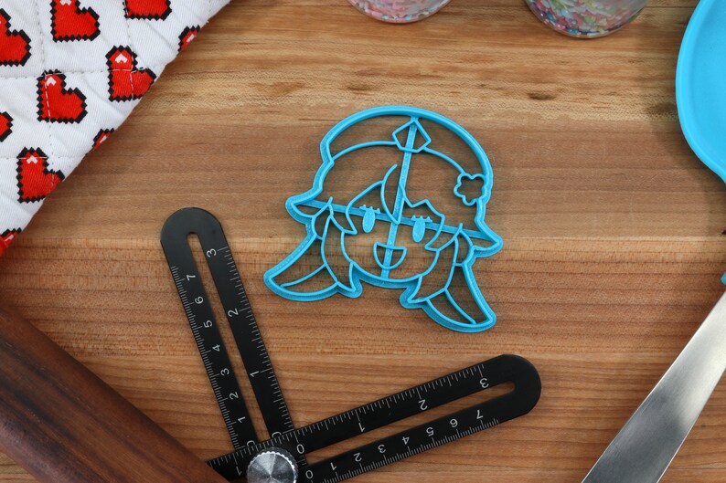 May include: A blue plastic cookie cutter shaped like a cartoon character with a flower in their hair. The character has a smiling face with closed eyes and a small mouth.
