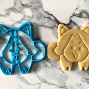 May include: A blue plastic cookie cutter shaped like a cartoon dog with a tongue sticking out, next to a light brown sugar cookie cut with the same shape.