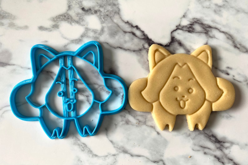 May include: A blue plastic cookie cutter shaped like a cartoon dog with a tongue sticking out, next to a light brown sugar cookie cut with the same shape.
