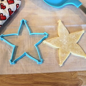 Pu&ograve; includere: Un biscotto a forma di stella accanto a un tagliabiscotti blu a forma di stella su carta da forno. Un guanto da forno a forma di cuore rosso e bianco &egrave; nell'angolo in alto a sinistra. Una spatola blu &egrave; nell'angolo in alto a destra.