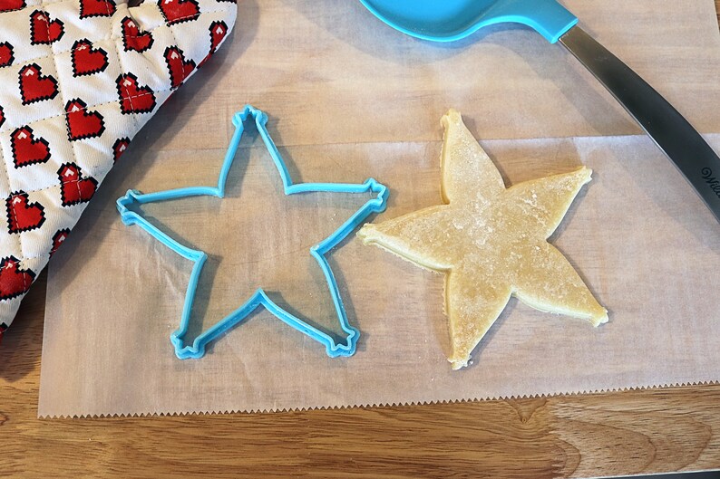 Pu&ograve; includere: Un biscotto a forma di stella accanto a un tagliabiscotti blu a forma di stella su carta da forno. Un guanto da forno a forma di cuore rosso e bianco &egrave; nell'angolo in alto a sinistra. Una spatola blu &egrave; nell'angolo in alto a destra.