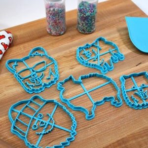 May include: Five blue plastic cookie cutters shaped like dogs, with different expressions and accessories. The cutters are on a wooden surface with a rolling pin and a spatula.
