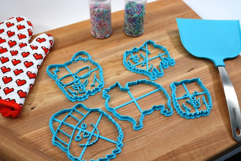 May include: Five blue plastic cookie cutters shaped like dogs, with different expressions and accessories. The cutters are on a wooden surface with a rolling pin and a spatula.