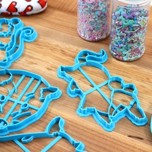 May include: A set of five blue plastic cookie cutters shaped like animals, including a squirrel, a pig, a unicorn, and two other animals. The cutters are on a wooden surface next to two jars of colorful sprinkles.