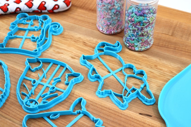 May include: A set of five blue plastic cookie cutters shaped like animals, including a squirrel, a pig, a unicorn, and two other animals. The cutters are on a wooden surface next to two jars of colorful sprinkles.