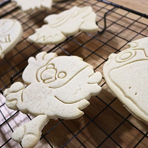 May include: Four plain sugar cookies on a black wire cooling rack. The cookies are shaped like cartoon characters and are ready to be decorated.