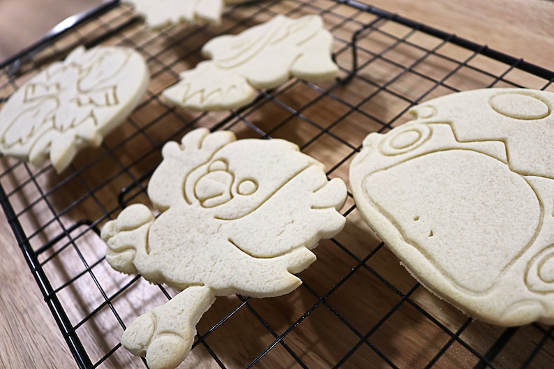May include: Four plain sugar cookies on a black wire cooling rack. The cookies are shaped like cartoon characters and are ready to be decorated.