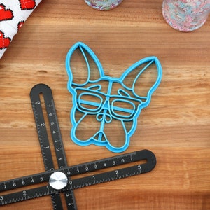 Puede incluir: Un cortador de galletas de pl&aacute;stico azul con forma de perro con gafas. El cortador de galletas est&aacute; sobre una superficie de madera.