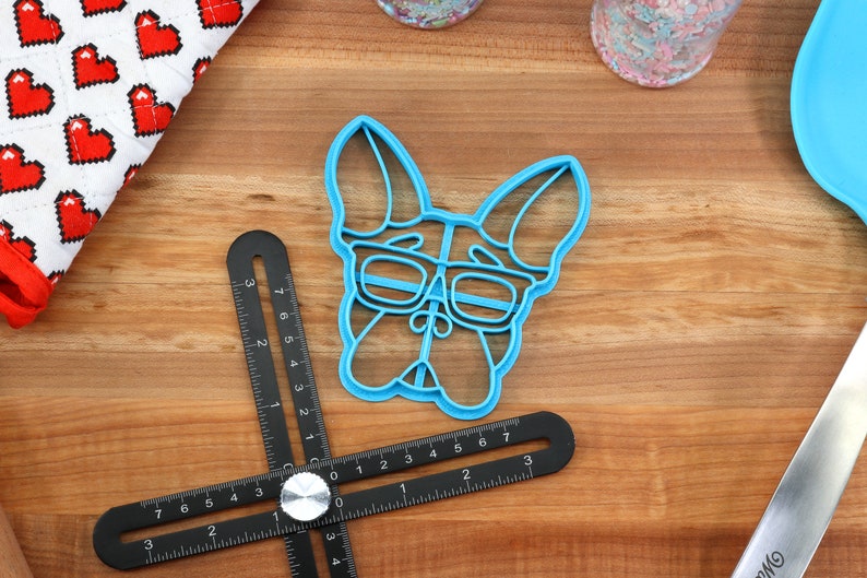 Puede incluir: Un cortador de galletas de pl&aacute;stico azul con forma de perro con gafas. El cortador de galletas est&aacute; sobre una superficie de madera.