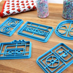 May include: A set of six blue plastic cookie cutters shaped like cassette tapes and a record player. The cutters are on a wooden surface.