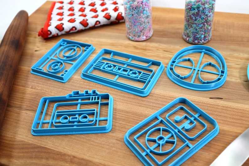 May include: A set of six blue plastic cookie cutters shaped like cassette tapes and a record player. The cutters are on a wooden surface.
