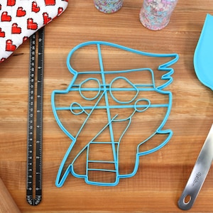 May include: A blue plastic cookie cutter shaped like a cartoon character with a hat and glasses. The cutter is on a wooden surface with a ruler, a rolling pin, and a blue spatula.