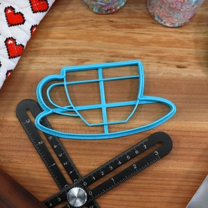 May include: A blue plastic cookie cutter shaped like a teacup and saucer. The cutter is on a wooden surface with a black ruler, a rolling pin, and a blue spatula.