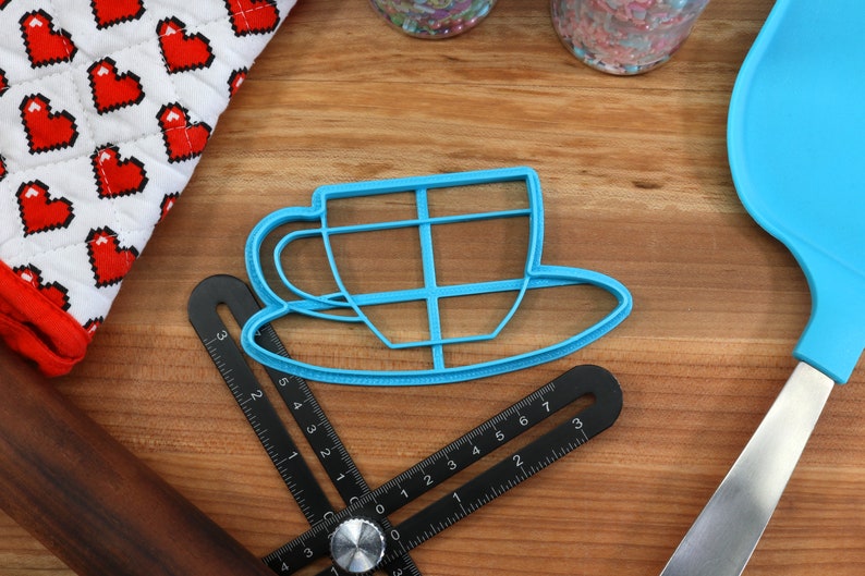 May include: A blue plastic cookie cutter shaped like a teacup and saucer. The cutter is on a wooden surface with a black ruler, a rolling pin, and a blue spatula.