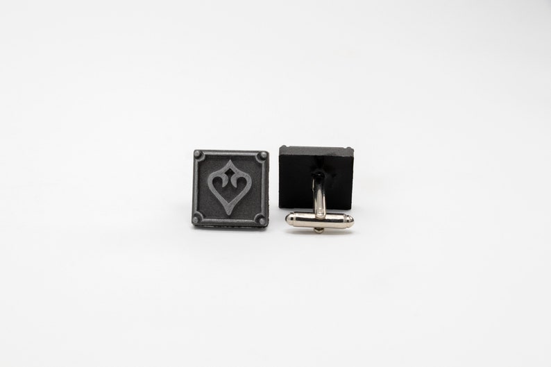 May include: A pair of silver cufflinks. One cufflink features a square design with a stylized heart in the center. The other cufflink is plain.