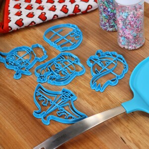 May include: A set of six blue plastic cookie cutters shaped like cartoon characters on a wooden surface. The cutters are next to a blue spatula and two jars of colorful sprinkles.