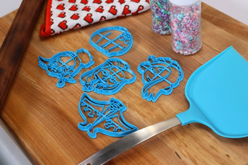 May include: A set of six blue plastic cookie cutters shaped like cartoon characters on a wooden surface. The cutters are next to a blue spatula and two jars of colorful sprinkles.