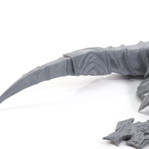Pode incluir: Close-up de uma garra de drag&atilde;o de pl&aacute;stico cinza e detalhada, com pontas afiadas. A garra est&aacute; parcialmente montada e pronta para ser pintada.