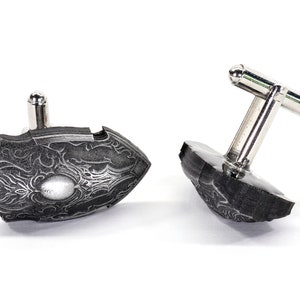 May include: A pair of silver cufflinks with a black, detailed, patterned design. The cufflinks are in the shape of a shield.