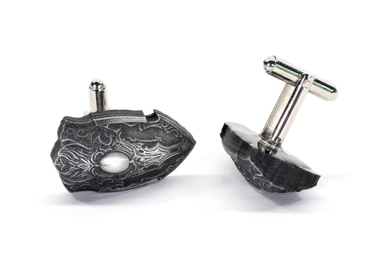 May include: A pair of silver cufflinks with a black, detailed, patterned design. The cufflinks are in the shape of a shield.
