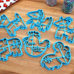 Puede incluir: Conjunto de siete cortadores de galletas de pl&aacute;stico azul con forma de varios animales, incluyendo un pollo, un rinoceronte, una cabra y un caballo. Los cortadores est&aacute;n sobre una superficie de madera.