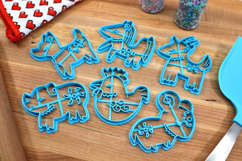 Puede incluir: Conjunto de siete cortadores de galletas de pl&aacute;stico azul con forma de varios animales, incluyendo un pollo, un rinoceronte, una cabra y un caballo. Los cortadores est&aacute;n sobre una superficie de madera.