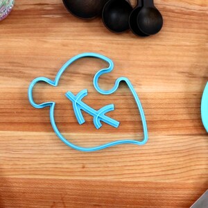 May include: A blue plastic cookie cutter shaped like a leaf with a small leaf design inside. The cutter is on a wooden surface.