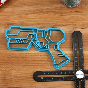 May include: A blue plastic cookie cutter shaped like a futuristic gun. The cookie cutter is on a wooden surface.