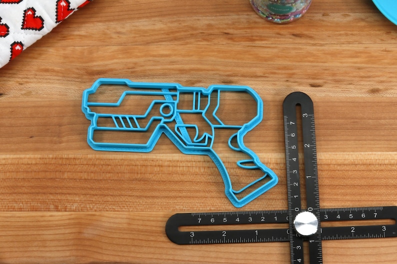 May include: A blue plastic cookie cutter shaped like a futuristic gun. The cookie cutter is on a wooden surface.