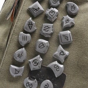 May include: A collection of 17 silver metal pins with various symbols and designs. The pins are attached to a green canvas bag.