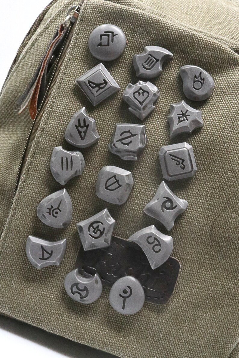May include: A collection of 17 silver metal pins with various symbols and designs. The pins are attached to a green canvas bag.