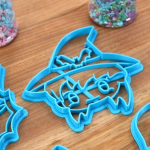 May include: A set of three blue plastic cookie cutters shaped like a witch, a bat, and a skull. The cutters are on a wooden surface.