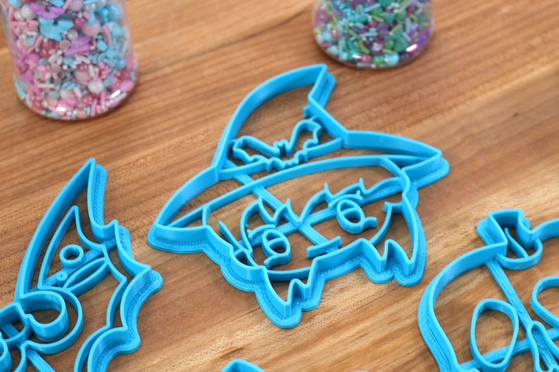 May include: A set of three blue plastic cookie cutters shaped like a witch, a bat, and a skull. The cutters are on a wooden surface.
