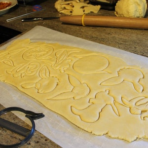 May include: A sheet of rolled out cookie dough with various shapes cut out using cookie cutters. The shapes include bunnies, flowers, and a circle. The cookie cutters are visible in the foreground.