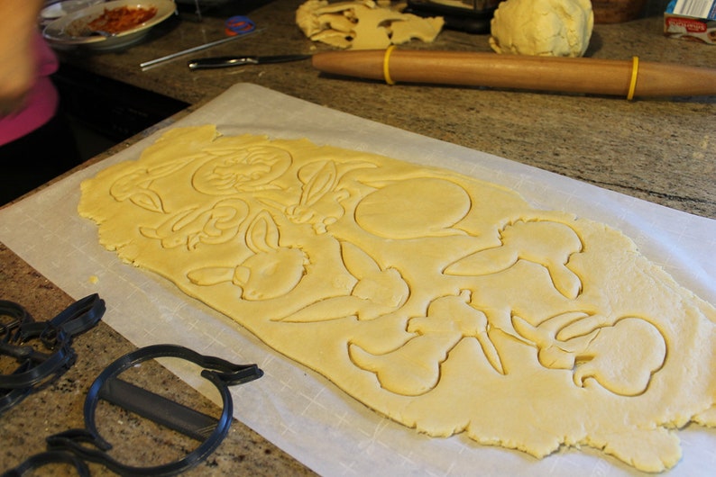 May include: A sheet of rolled out cookie dough with various shapes cut out using cookie cutters. The shapes include bunnies, flowers, and a circle. The cookie cutters are visible in the foreground.