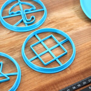 May include: A collection of light blue cookie cutters in various shapes, including an umbrella, a star, and a square grid, arranged on a wooden surface. A blue spatula and a ruler are also visible. The cookie cutters are designed for baking.