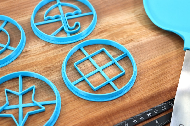 May include: A collection of light blue cookie cutters in various shapes, including an umbrella, a star, and a square grid, arranged on a wooden surface. A blue spatula and a ruler are also visible. The cookie cutters are designed for baking.