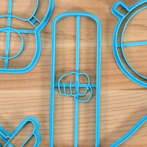 May include: A set of five blue plastic cookie cutters shaped like a dumbbell, a fist, a circle with a cross in the center, and two other shapes.