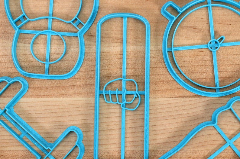 May include: A set of five blue plastic cookie cutters shaped like a dumbbell, a fist, a circle with a cross in the center, and two other shapes.