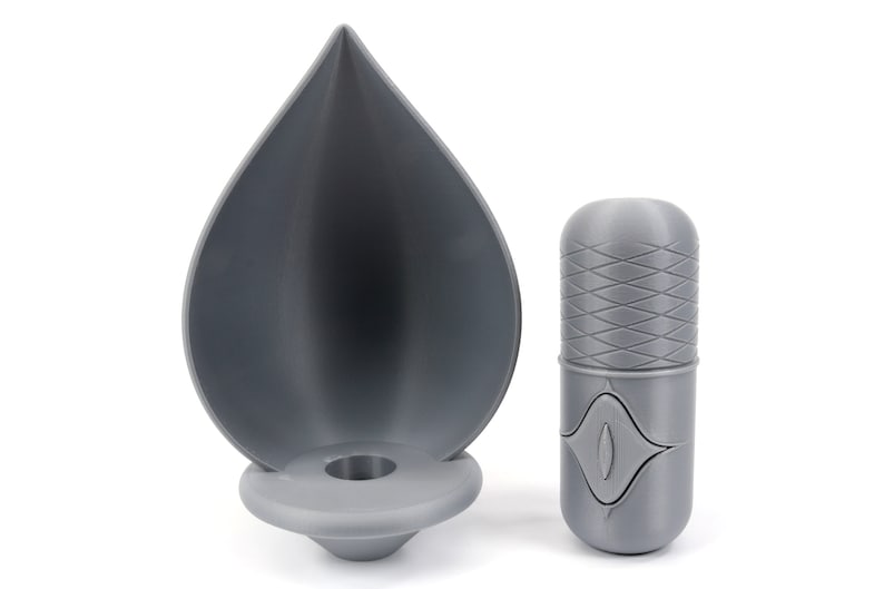 May include: A gray 3D printed candle holder shaped like a teardrop with a round base and a small hole in the center. Next to it is a gray 3D printed cylindrical container with a textured pattern and a small eye-shaped design.
